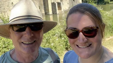 Family picture Woman and man smile at the camera, both wearing sunglasses. You can see the botom of a castle behind them and some greenery. The man is wearing a sunhat.
