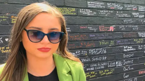 Tom Dunn/BBC Grace Merritt, 22, who has long straight light brown hair and is wearing sunglasses, a green blazer and a black top, stands in front of a black wall covered in messages written in different colours.