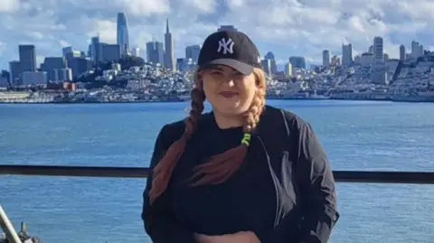 Jill Bunyan June wearing a black NY baseball cap and black clothing. Behind her is water and then the skyline of San Francisco.