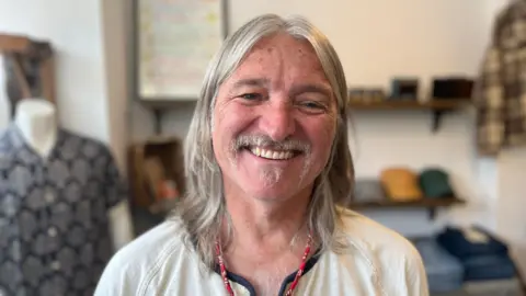 Lucinda Adam/BBC Clive has shoulder-length grey hair and a goatee beard. He wears a cream top.