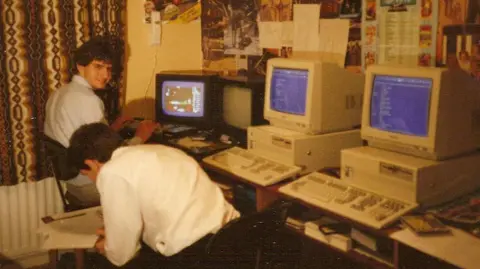 Handout An archive photograph of the twins as young men, wearing dark trousers and light coloured collared shirts. They are sitting at a desk in front of four large computers, which look outdated and oversized. On them are coding screens and prototype gaming software. The room looks retro with patterned curtains and lots of posters on the wall.