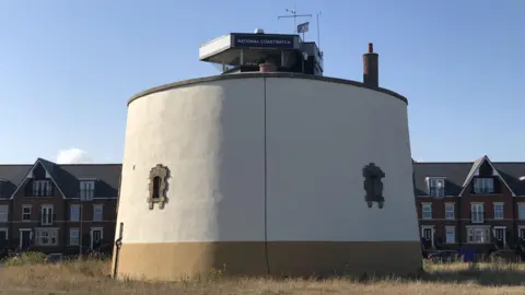 The round grey exterior of Martello Tower P in Felixstowe featuring small windows reinforced with bricks and a National Coastwatch lookout on top 