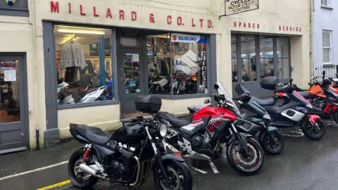 BBC Millards bike shop from the exterior. A creamy yellow building with motorbikes in front and a red sign which says Millards & Co Ltd.