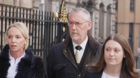 Ian Harvey, with short grey hair and a short grey beard, walks towards Liverpool Town Hall with a serious expression, flanked by two women, one with blonde hair and one with brown hair