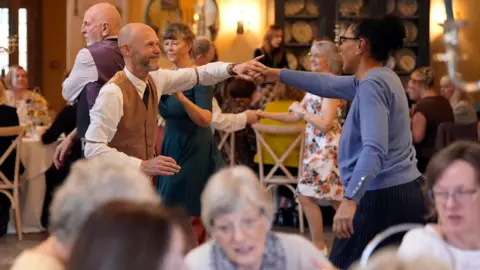 PA Media A man and a woman swing dance in the centre of a dining room, surrounded by tables of people enjoying afternoon tea. Other people are stood up, dancing behind them.