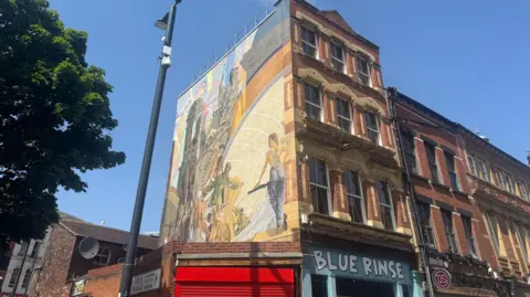 Dan Bater/BBC A building in Leeds which has a mural on the side which is called Cornucopia. The building is red brick and has shops on the ground floor, including one called Blue Rinse. 