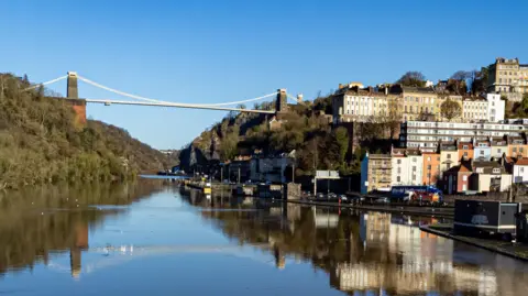 Clifton Suspension Bridge and its surrounding area of Bristol on a sunny day. The bridge can be seen in the reflection on the River Avon. To the right, houses staggered up a hill can be seen glowing in the winter sunlight.