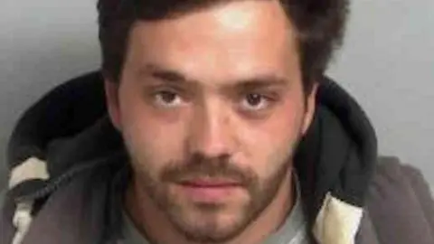 Essex Police Charles Benson-Clark wearing a grey crew-neck shirt layered under a dark jacket, seated against a plain light grey background. He has black hair.