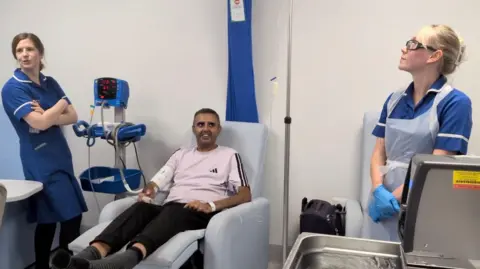 Sheffield Teaching Hospitals NHS Foundation Trust A man is sitting in a chair with his legs raised in a hospital treatment room. There are two nurses in the room on either side of him. They are preparing to give him an intravenous treatment.