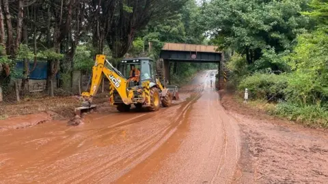 A large yellow digger removing silt and mud from the road. It has cleared a path through the banks of orangey-brown mud which still cover the road.