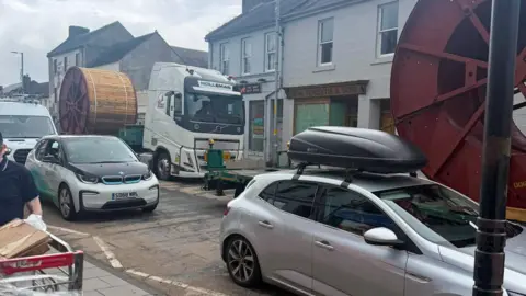 The main road through Sanquhar clogged up with traffic, we can see cars in the foreground and a lorry with a large, round, wooden cog on the back in the background. The vehicles appear to be at a standstill.