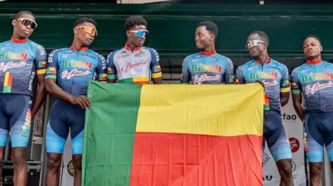 Six male cyclists from Benin wear blue cycling gear with the word Benin on the chest and stand in a line to pose behind a flag of the West African country