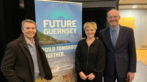 The three leading members of Future Guernsey. On the right is Deputy Gavin St Pier, wearing a black suit and a blue and white dotted tie, over a blue shirt. Next to him is CEO Lindsey Freeman who is wearing a black zip up jacket. Next is a banner that says Future Guernsey with the background a picture of Guernsey's coast and a slogan which says 'Build together tomorrow'. On the far-left is John Hardie wearing a brown jacket. 