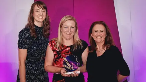 Mark Allen Group Milly poses with comedian Lucy Porter and an award sponsor, and holds her award. It is circular and purple/pink, with two ears of golden corn. Milly wears a black dress with a large red flower on one side.