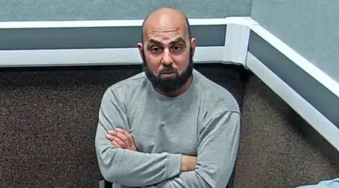 Amar Hussein is interviewed in a police station. He is largely bald with a trimmed black beard. He wears a light grey tracksuit and is sitting with his arms folded across his chest