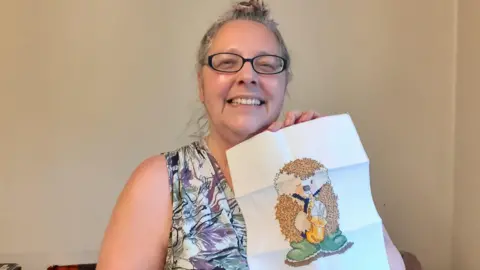Rose Lofas Molly Lofas, who has glasses and tied-up grey hair, smiles as she holds up a cross-stitch of a hedgehog in a blue tie and green wellington boots playing a saxophone