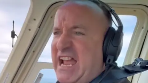Dave Fishwick Dave Fishwick, wearing a headset and sitting in the cockpit of his helicopter, is narrating what he can see out of the window.