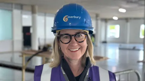 Caoimhe Keogan is standing in the middle of a room which is currently under construction. She is wearing a blue hard hat and a purple Hi-Vis jacket.