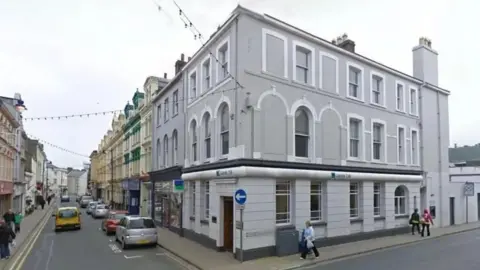 A grey, Victorian-style building on a street corner in Ramsey. Cars can be seen driving along and parked on Parliament Street.