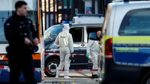 In focus, two police in white hazmat suits walk by a black car. In the foreground, obscuring the car and one policeman, is a police van and a uniformed officer 