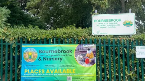 A large temporary sign is attached to green railings advertising places available at Bournebrook Church of England Primary School & Nursery. On the top right there is a permanent white sign with green writing for Bournebrook Church of England (voluntarily controlled) Primary School. Behind the railings is a hedge and some trees.
