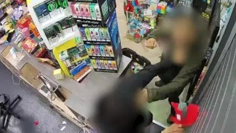 LDRS A CCTV image shows a shop assistant being assaulted inside a convenience store in Liverpool. A man whose face is blurred can be seen throwing a punch over the counter at the person working behind it. 