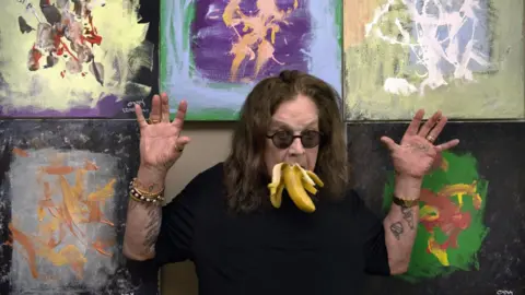 Kamil Szkopik A man is wearing a black top and sunglasses and is standing in front of five painted canvases. He is holding his hands in the air and has a banana in his mouth.