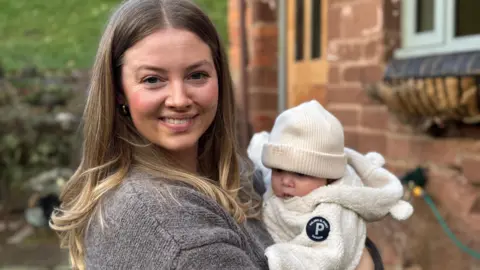 Baby Penny in her mother's arms. She wears a cream hat and cream jacket while her mum has long dark blonde hair and a grey top.