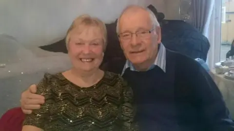 Mavis and Dennis are sitting together, and Dennis has his arm around his wife. He had short white hair and glasses. Mavis has short blonde hair.