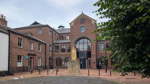 To the right in the foreground there is part of a green tree. In the centre there is a plinth with a yellowing statue. In the background there is a red brick building with large glass windows. At the top of the building there is a crest.
