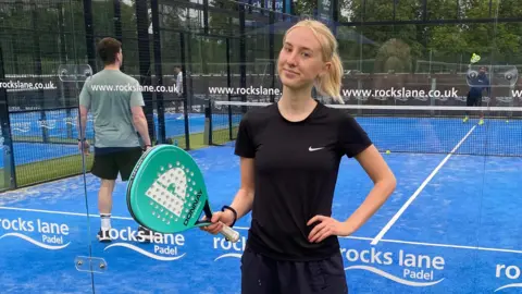 Rosie is standing in front of a blue astro padel court. She is wearing a black t-shite and matching tracksuit bottoms and we can only see her from the waist up. She is holding a turquoise padel racket in one hand and the other hand is on her hip. Rosie has shot blonde hair, which has been tied back into a low ponytail. She is smiling into the camera with no teeth. Behind her are two men playing on the court, who can be seen through the glass walls. There is white branding written on the glass. 