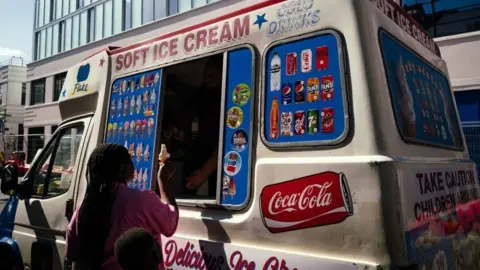 A mother and child stand next to an ice cream van while the mother buys an ice cream cone