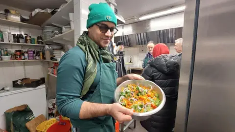 Sâmân Jamshidifard Sâmân Jamshidifard in a kitchen, holding a bowl with a vegetable salad in it. On the shelves behind him, there is cutlery, crockery and various cooking ingredients. Three men and a woman are also in the kitchen, having a conversation behind him.
