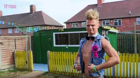 Tomasz in his garden wearing a denim gilt with no shirt on. He has Jedward-like hair and has a bow tie on.