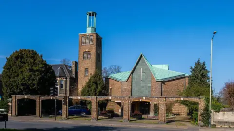 A 1950s church is shown under clear blue skies with trees to its sides. It has a faded blue roof and a cross on its exterior with a tall tower and arched walls.
