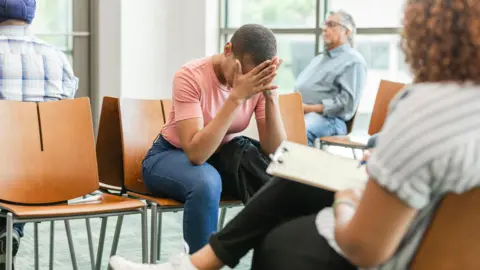 A waiting room. One woman sits with her head in her hands, while a man rests his head back on his seat. In the foreground there is a woman writing notes on a clipboard.
