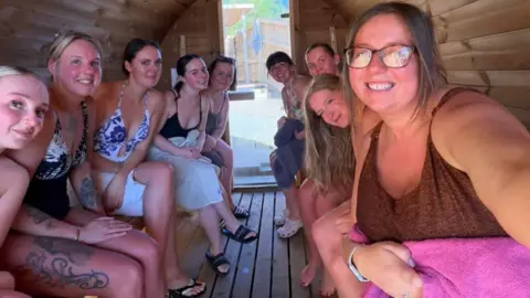 Izzy Taylor A group of women sit together in a sauna, looking at the camera, wearing swimwear and towels