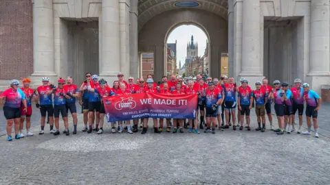 RBVE A group of people, in red and blue cycling gear, standing in front of a large building. They are holding a red and blue banner with the words "we made it".