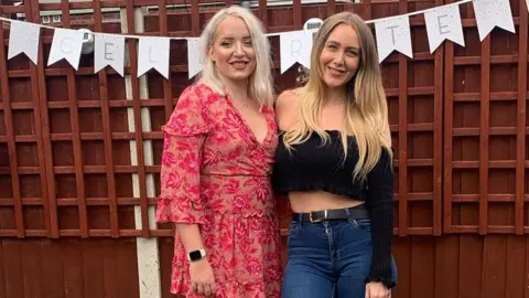 Ashleigh Charlesworth Ashleigh and April stand next to each other with their arms around each other. They are smiling to the camera in front of a fence with a banner hanging on it. Ashleigh has shoulder length white hair and April has long blonde hair.