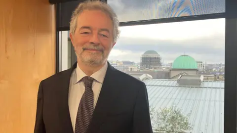 Austin Hughes is standing in a room in an office. Roofs can be seen from a window behind him. He's wearing a black suit with a white shirt and a black and white tie.