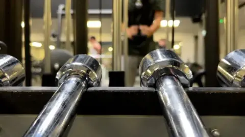 Getty Images Two silver dumbbells in foreground in a rack with unidentifyable people in the background using gym equipment