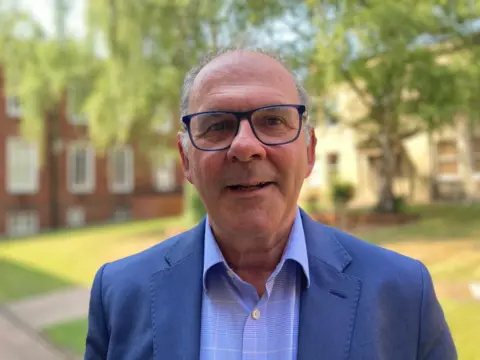 Prof Neal Juster is wearing a blue suit and a lighter blue shirt, he has dark framed glasses and grey hair. He is standing in a lawned courtyard with a tree in the centre.