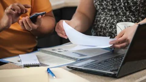 A close up of two women's hands while they are sat at a desk. There is an open laptop and a notebook, pen and mug on teh table. One of the women is typing into her phone while the other is holding paper documents.