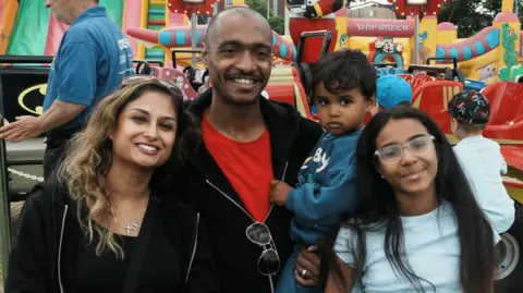 A woman, man and two children are smiling at the camera. The man is holding a small boy in his arms and a young girl is standing beside them.