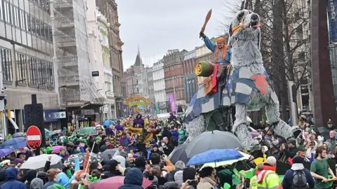 Pacemaker A packed city centre street has huge numbers of crowds lining the streets to view a parade. A number of figures in fancy dress make their way through the crowd