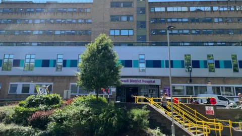Outside of Yeovil district hospital's main entrance, with yellow steps and green bushes. The building is brown bricks