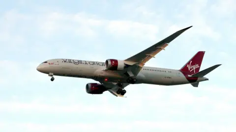 A large white passenger plane in flight. It carries red Virgin Atlantic branding.