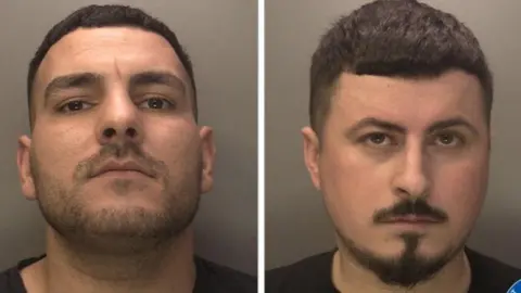 Two mugshots next to each other of two men. They both have short dark hair. The man on the left has stubble and is holding his chin up while staring into the camera. The man on the right has black moustache and goatee, he is staring into the camera as well.