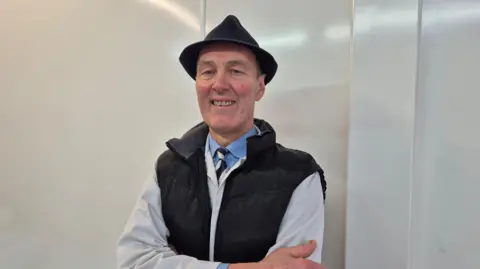 James Baillie standing in his butcher's shop. He is wearing his work clothes and smiling for the camera.