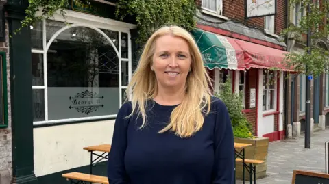 A woman with blonde hair smiling standing outside a pub with an Italian restaurant in the background
